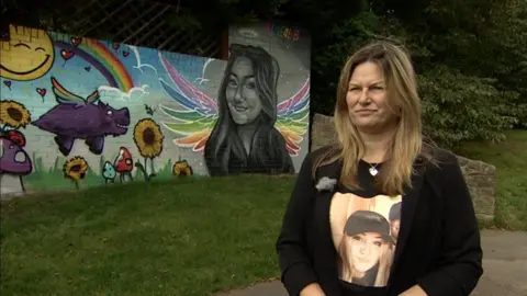 A blonde woman stands in front of a multi-coloured mural on a wall, which features a painting of Liberty in black and white, with rainbow-coloured angel wings. Other features of the mural include a sun, rainbow, sunflowers, and a purple flying animal with multi-colour wings. The woman wears a black t-shirt with her daughter's face printed on it.