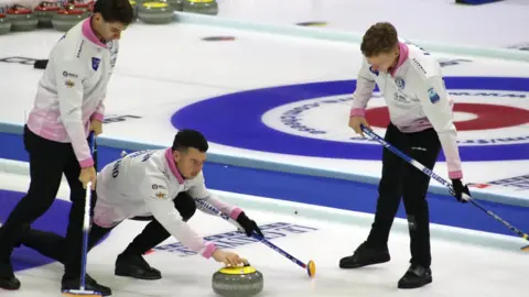 British Curling Team Mouat players Grant Hardie, Hammy McMillan and Bobby Lammie in action in Finland, as Hammy prepares to throw the curling stone and Grant and Bobby stand either side preparing to sweep