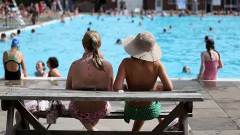 The back of two women sat at a wooden bench, looking out over a busy outdoor swimming pool. One of them is wearing a hat and it is very sunny.