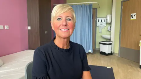 Person with short blonde hair wearing a dark top, seated in a clinical examination room with a hospital bed, privacy curtain, sink, and medical fixtures visible in the background.
