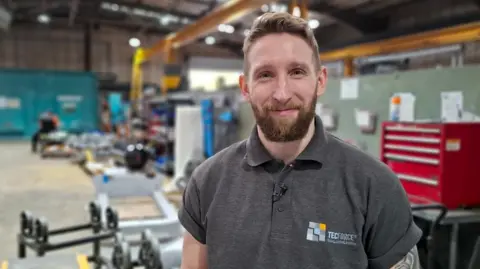 Peter is wearing a Tecforce T-shirt and is stood on the factory floor smiling at the camera. He has a styled hair do quiff, with a short beard.