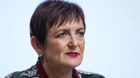 Angela Constance, a woman with short purple hair. She is wearing dark pink lipstick and a patterned top.