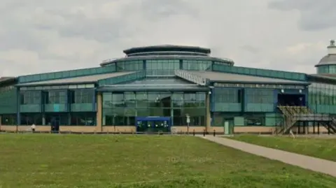 Google Exterior of the shopping centre- a green glass building with grass in the front.
