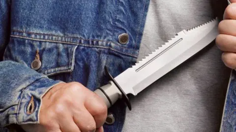 Getty Images A stock image of a person holding a large silver and grey zombie knife. The picture is a close up of their hands and torso, and they are wearing a grey top and blue denim jacket. 