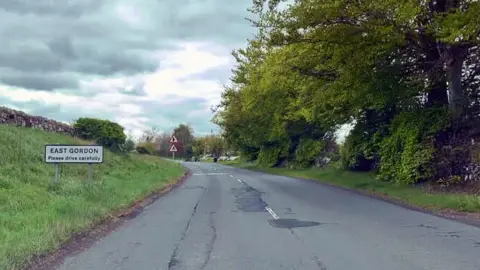 A general view of the A6105 near East Gordon. There is a road sign on the left and trees lining the road on the right.