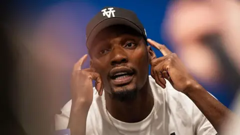 US track and field sprinter, Fred Kerley, talks with the press ahead of the Enhanced Games. He points to his face with his hands, mid-conversation.