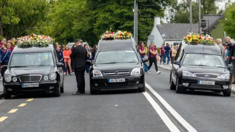 PA Media Three funeral hearses driving on a road with flowers on top of them. Crowds of people are behind it.