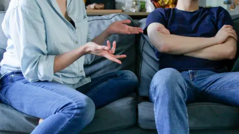 Getty Images Two people sitting on a blue sofa, looking like they are in an argument. The woman has her hands out, and is wearing blue jeans and a blue shirt, the man, next to her is also wearing blue jeans and a blue T-shirt, you can only see part of their legs and body.