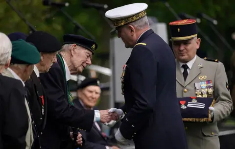Chris Redston A line of former World War Two veterans stand in military uniform. One of the men, wearing a military cap and jacket, shakes the hand of a man in front of him.