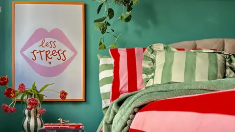 Rachel Verney A bedroom with green walls has plants hanging from a shelf and a bed is made up with red-and-pink striped bedding. A picture frame that says 'Less Stress' is hanging up on the left side of the bed.
