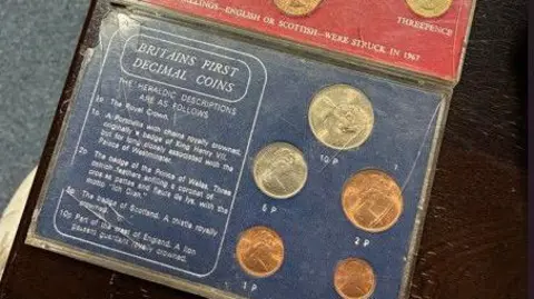 A clear box contains a selection of decimal coins with brief descriptions from 1969. Slightly visible are the set of coins above it which are pre-decimal currency in the UK. 
