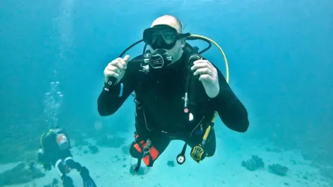 Rob Mason A man is wearing a full scuba diving suit and equipment and is underwater in clear blue water.