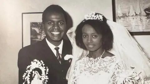 Fumi Stephenson Mr and Mrs Stephenson in a black and white wedding picture. They are inside, with framed pictures behind them, and are dressed in their wedding clothes. Dr Stephenson is wearing a dark suit and tie with a flower in his button hole, while Mrs Stephenson wears an ornate white lace dress, a large fluffy veil and a tiara in her hair. 