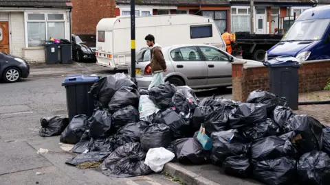 A large pile of black bin bags on the side of a residential street in Birmingham. A pedestrian is walking past the bags, and there are various vehicles parked on both sides of the road.