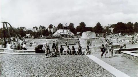 Cheltenham Lido This black-and-white image captures Cheltenham Lido in 1981. A slide is visible to the left, while the poolside is bustling with activity, with children in swimsuits relaxing on the surrounding grass.