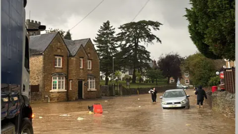 Droneovermk/Instagram A picture of a road in Lavendon that has been hit by heavy flooding. A car can be seen partially submerged as people wade through the water. 