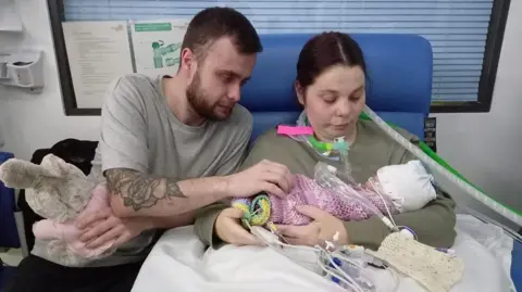 Irwin Mitchell Adin Humphries sits on a chair in a hospital ward with his partner Chelsea Wootton as he holds their daughter Ava-Lee, who is hooked up to medical equipment.