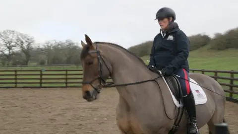 Sara Bates rides Tango, a brown horse, around a sandy dressage arena, surrounded by wooden fence. She is wearing black riding boots, blue and red jodhpurs, a blue zip-up fleece and black riding helmet. 