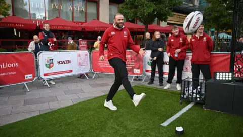 Getty Images Jamie Roberts wearing a Lions jersey kicking a rugby ball at a rugby promotion event. There is a barrier around the artificial turf with people watching from the sides. The Ivy Asia restaurant is seen in the background on the left.