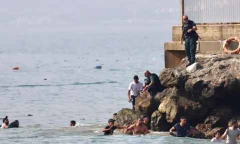 Men swimming as police officer standing on rocks holds arm of man in white t-shirt