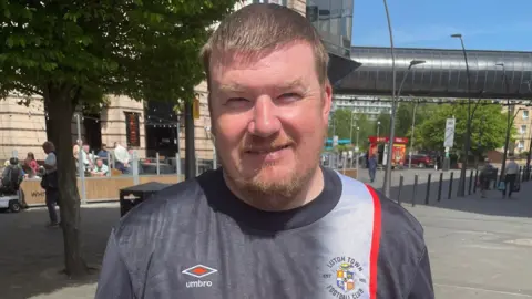 James Burridge/BBC Kevin stands in the square in front of trees and a cafe. He wears a grey Luton town t-shirt with a red stripe and Umbro logo. He has short blonde hair, and a short beard and moustache.