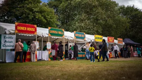 Leeds Road Festival People buy food from stalls on a field: Doner house, Samoa Wala, Biryani Hut, Jalebi Baby