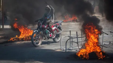  EPA-EFE/REX/Shutterstock A man rides a motorcycle as tyres burn in Port-au-Prince, Haiti. Photo: March 2024