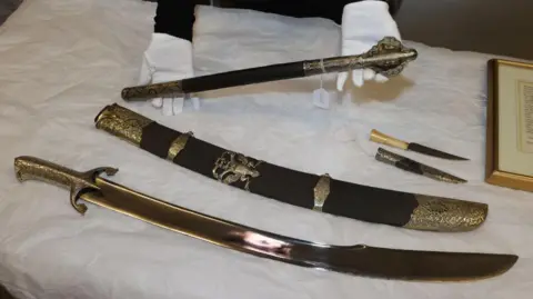 Old weapons laid out on a table - a scimitar (sword/sabre) and scabbard, a mace, and a dagger and sheath.
