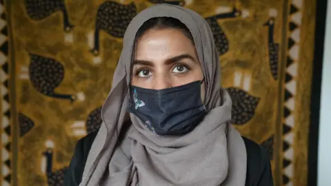 Bella is wearing a head scarf and face mask in front of a wall hanging