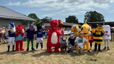 John Fairhall/BBC A number of mascots in a variety of furry suits stand in the grand ring. They include Crazee and Bluey from Ipswich Town football club, a bumble bee, a monkey and a red dinosaur
