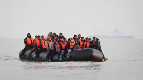 PA Media Migrants sit in an inflatable boat as they travel across the English Channel. They are wearing orange life jackets.