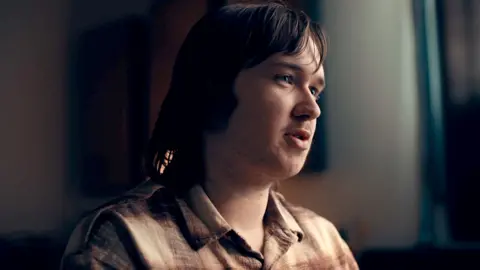 Calum, a young man with long brown hair and slight facial hair, wearing a brown checked shirt, pictured in close-up looking to the right of the camera in a darkened room with the curtains behind him drawn, stark light falling across his face from the right of the image.