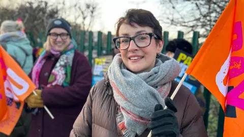 A woman with short dark hair and black-rimmed glasses smiles as she holds an orange NEU flag on a picket line outside green college gates. She is wearing winter clothes including a padded coat and a big grey scarf. A fellow member of staff can be seen holding a flag in the background.