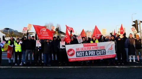 Ross Quinn/Supplied A small group of protesters holding flags and placards pushing for a pay rise. They are stood at the side of a road and dressed warmly in puffer jackets, jeans and woolly hats