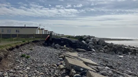 Coastline at Walney Island. A grassed area with some static caravans can be seen on the left, while a beach area is covered in pebbles. Large rocks are positioned in front of a stretch of road to provide a defence against tides.