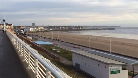 A view of Bridlington's South beach, with a promenade and a large sandy beach, with the sea to the right. In the background, large buildings and a harbour, with the coast sweeping around to a headland.