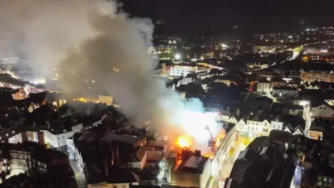 UKNIP A drone shot of smoke coming from a building's roof which is on fire. There are two aerial platforms above the fire.