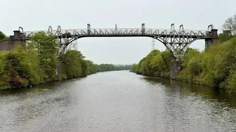 A metal bridge straddles two wooded banks over a large river on a clear day.