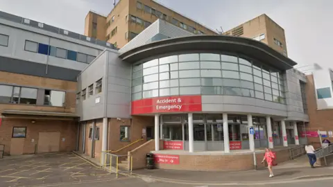 Google Exterior of Yeovil District Hospital's A&E department. The building is a mixture of brown bricks and grey panels with A&E sign. There are a few people walking around near the entrance. 