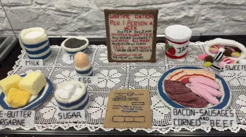 Rheged A knitted scene representing rationing. It shows several items of food such as butter, sugar, milk, meat and sweets laid out on plates on a table, with a ration card and a board stating how much each person is entitled to.
