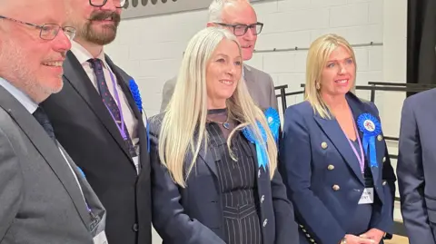 Paul Moseley/BBC Three men and two women wearing suits and blue rosettes. Kay Mason Billig is the central focus of the image. She has a navy jacket over a black pantsuit with white pinstripes.