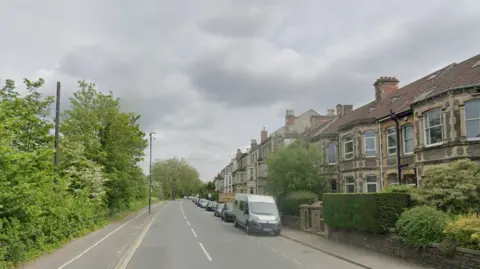 Google Coronation Road in Southville, Bristol. Houses are lining the street to the right, and trees and nature to the left. Cars and vans can be seen parked on the side of the road. 