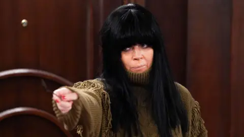 BBC Claudia Winkleman played by Anna Friel, who is wearing a brown fringed jumper and a black wig with a blunt fringe.