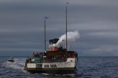 Margaret Howie The Waverley paddle steamer sails across a choppy sea under a grey sky. White steam billows from the vessel’s black-and-white funnels, and passengers line the decks. Two tall masts display maritime flags, including Scottish saltire flags. A small boat follows behind on the left, while faint outlines of distant hills or islands are visible on the horizon.