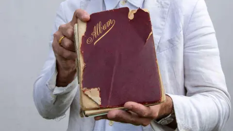 Hanson Auctioneers An old book with a worn, red cover and gold writing saying 'Album' being held by a man wearing a white overcoat.