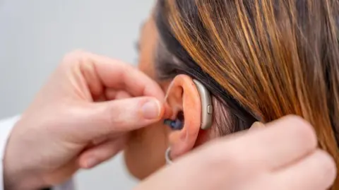 A close-up of a hand which is fitting a hearing aid to a woman's ear. The picture is taken from behind the woman's head. She has dyed brown hair.