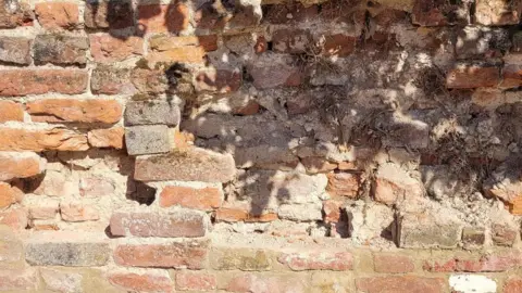 Andy Massey A close up of the damaged, weathered brick wall. The masonry is aged, with areas of erosion, missing mortar, and deteriorated brickwork.