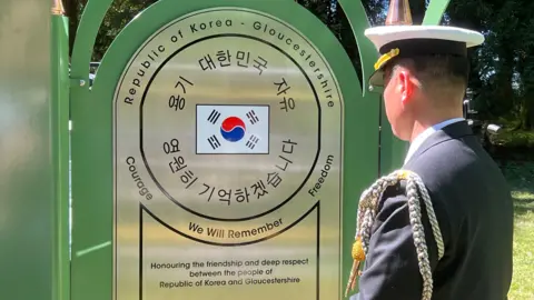 A man wearing military uniform with a white cap and ropes across his shoulder. He is looking at the memorial, which is a large metal rounded plaque on a metal green frame. There is a Korean flag in the centre, around which is says 'Courage, We will Remember, Freedom. Honouring the friendship and deep respect between the people of Republic of Korea and Gloucestershire'.