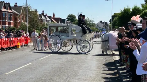 People lining street applaud as carriage with coffin turns into church