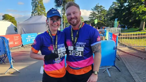 Collect/PA Real Life Smith and her husband are standing with their arms around each other smiling. Smith has a blue cap on, with a blue, pink and orange t-shirt. She has a breathing strip on her nose. Her husband is wearing the same top. They both have a black half marathon medal around their necks